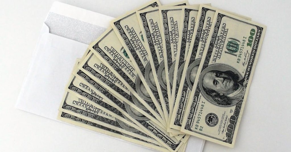 Fan of US $100 bills partially out of a white envelope on a white background.